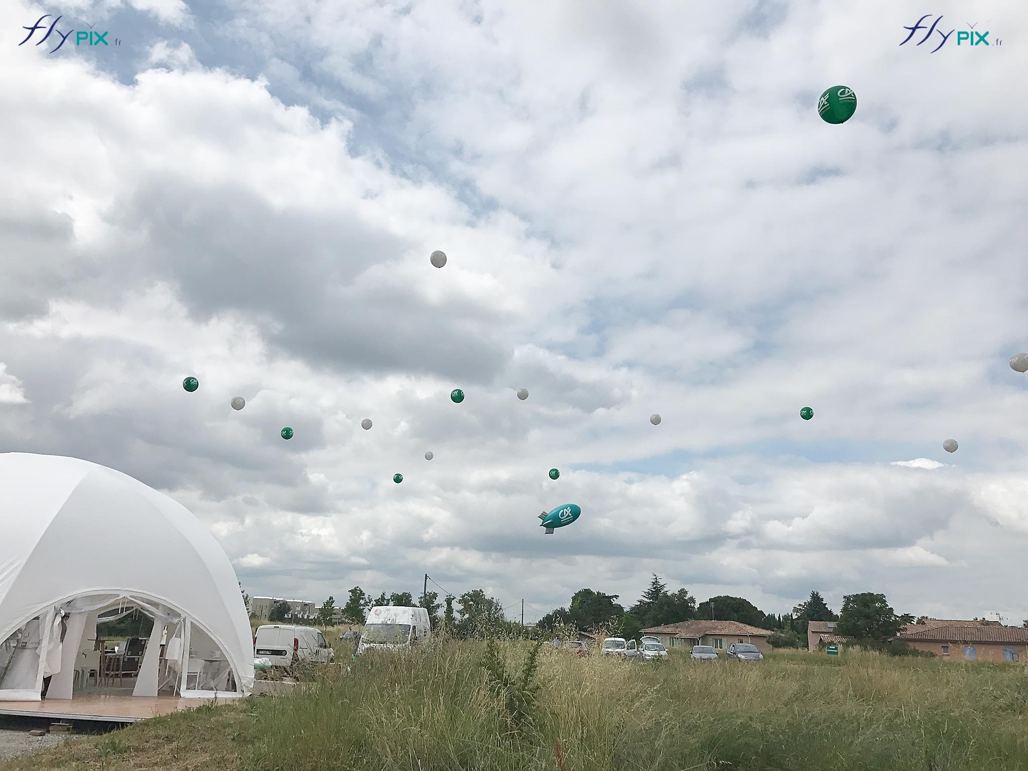 Plusieurs ballons personnalisés gonflés à l'hélium en vol au dessus d'un stand pendant un salon en extérieur, un jour de beau temps.